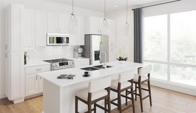 kitchen with appliances with stainless steel finishes, pendant lighting, light hardwood / wood-style flooring, white cabinets, and an island with sink