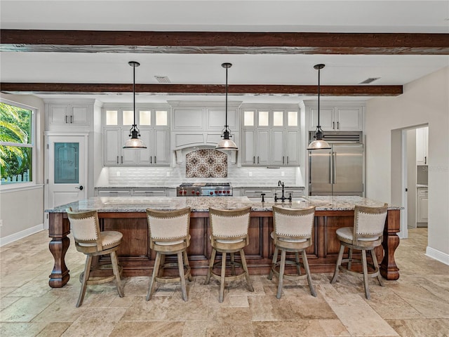 kitchen featuring a kitchen bar, built in fridge, an island with sink, decorative light fixtures, and light stone counters