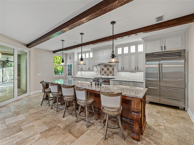 kitchen with beam ceiling, light stone counters, decorative light fixtures, a kitchen island with sink, and high end appliances