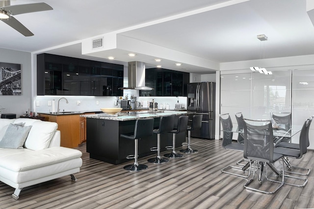 kitchen with a center island, wall chimney exhaust hood, stainless steel fridge with ice dispenser, light stone counters, and a breakfast bar area