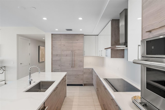 kitchen with black electric stovetop, stainless steel oven, wall chimney range hood, sink, and white cabinetry