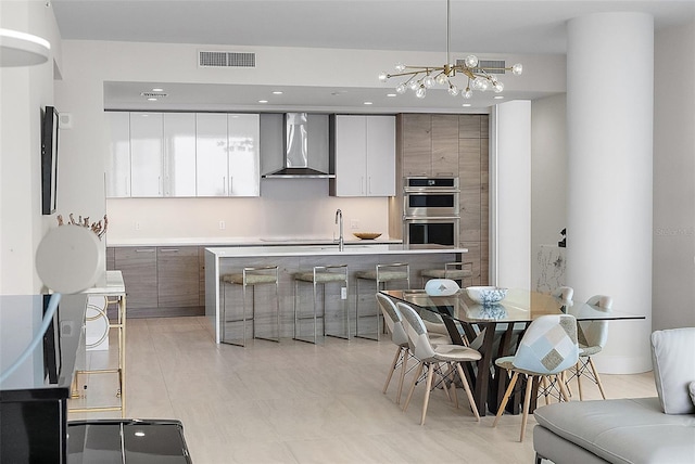 kitchen with pendant lighting, white cabinets, wall chimney range hood, double oven, and a chandelier