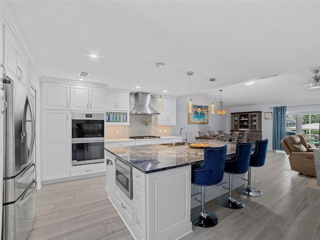 kitchen featuring dark stone counters, a center island with sink, white cabinets, wall chimney range hood, and appliances with stainless steel finishes