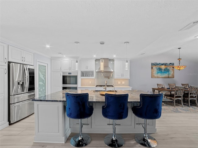 kitchen featuring wall chimney exhaust hood, stainless steel appliances, decorative light fixtures, a center island with sink, and white cabinetry