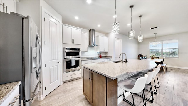 kitchen with white cabinetry, wall chimney range hood, decorative light fixtures, a kitchen island with sink, and appliances with stainless steel finishes