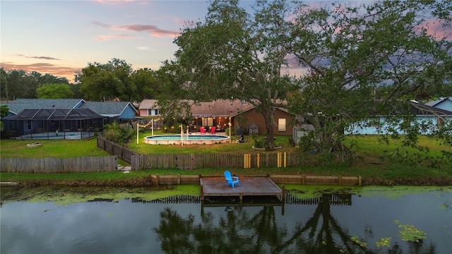 back house at dusk with glass enclosure, a water view, a yard, and a fenced in pool