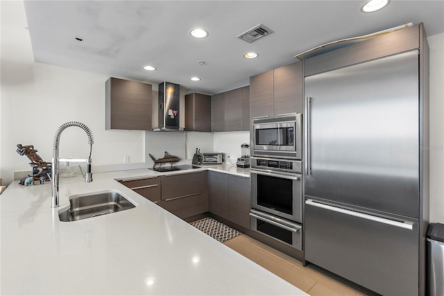 kitchen featuring dark brown cabinets, built in appliances, wall chimney exhaust hood, and sink