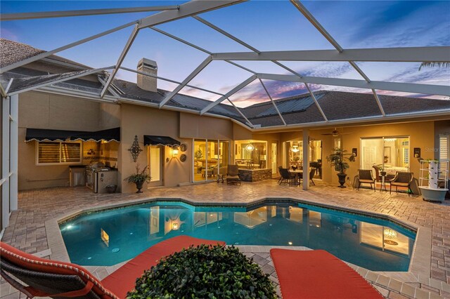 pool at dusk featuring a patio, an outdoor hangout area, exterior kitchen, and glass enclosure