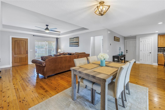 dining room with ceiling fan, a textured ceiling, a raised ceiling, and light wood-type flooring