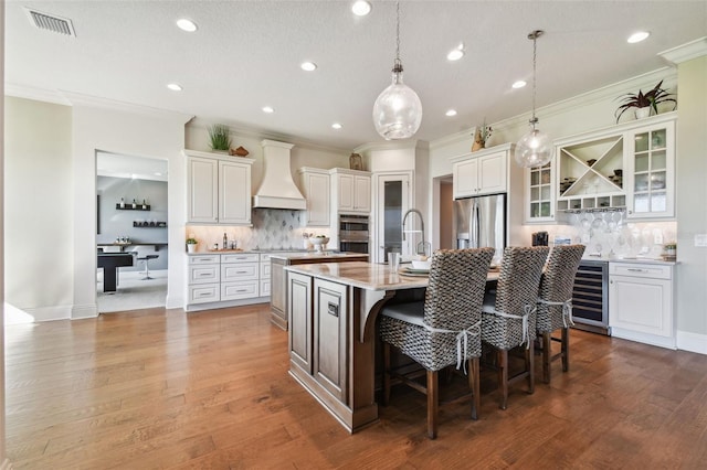 kitchen featuring an island with sink, a breakfast bar, white cabinets, custom range hood, and appliances with stainless steel finishes