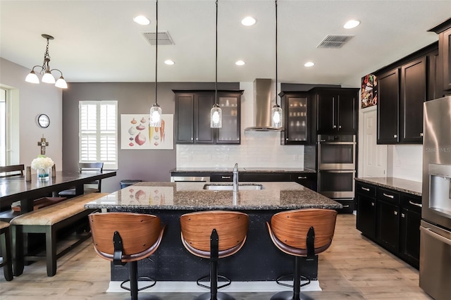 kitchen featuring pendant lighting, a kitchen island with sink, wall chimney range hood, sink, and stainless steel appliances