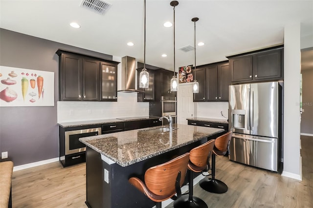 kitchen with sink, stainless steel appliances, wall chimney range hood, an island with sink, and pendant lighting