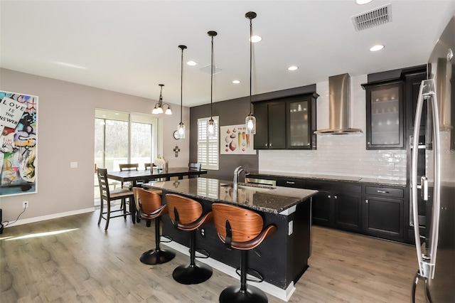 kitchen featuring a breakfast bar, dark stone counters, a center island with sink, sink, and wall chimney exhaust hood