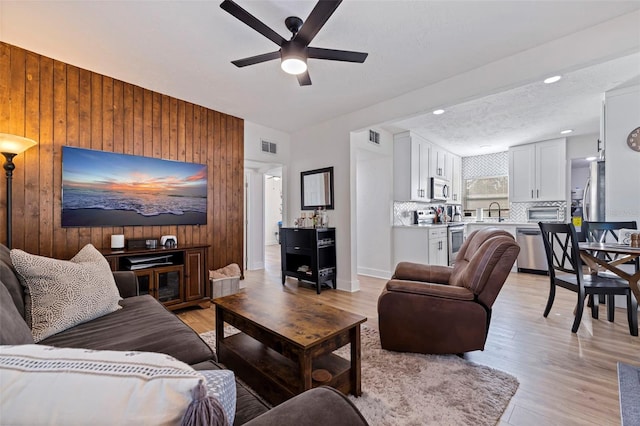 living room with a textured ceiling, ceiling fan, wooden walls, sink, and light hardwood / wood-style flooring