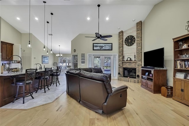 living room featuring high vaulted ceiling, french doors, ceiling fan with notable chandelier, a brick fireplace, and light wood-type flooring