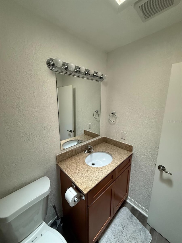 half bathroom featuring a textured wall, toilet, wood finished floors, vanity, and visible vents