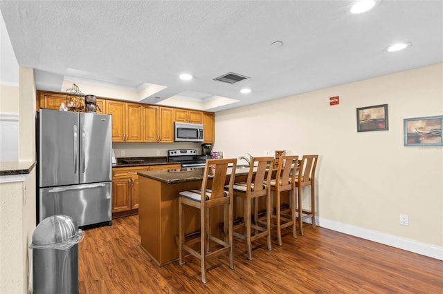 kitchen with appliances with stainless steel finishes, dark hardwood / wood-style flooring, a kitchen breakfast bar, a raised ceiling, and crown molding