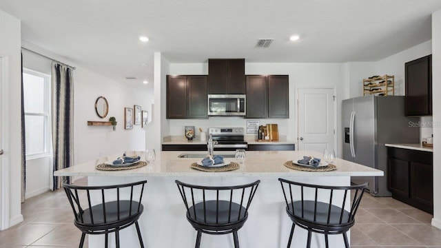 kitchen with dark brown cabinets, stainless steel appliances, light tile patterned floors, and a center island with sink