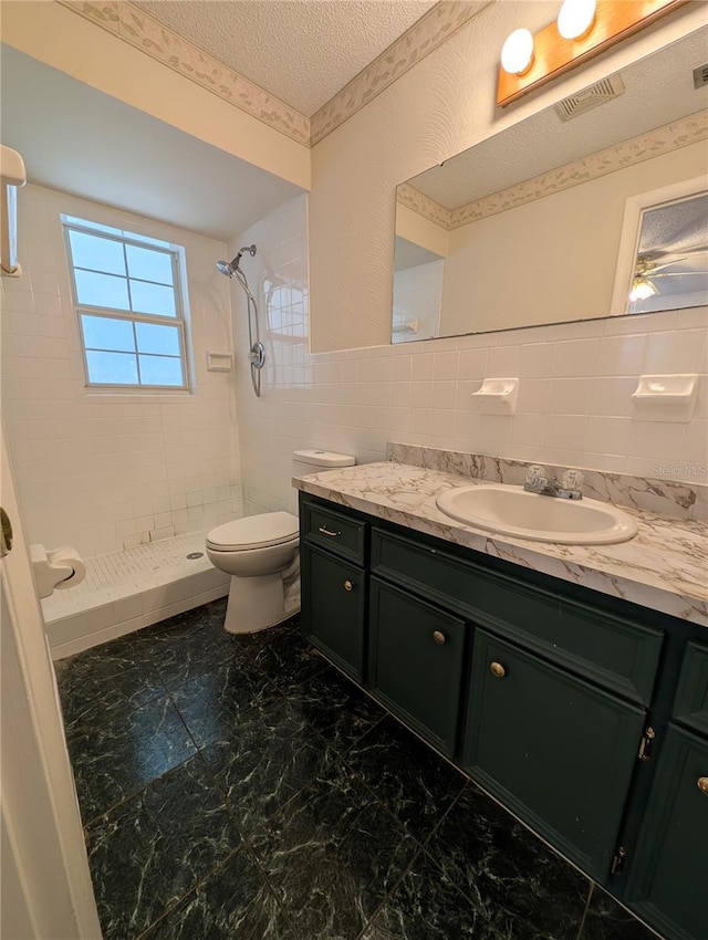 bathroom featuring a textured ceiling, toilet, tiled shower, vanity, and tile walls