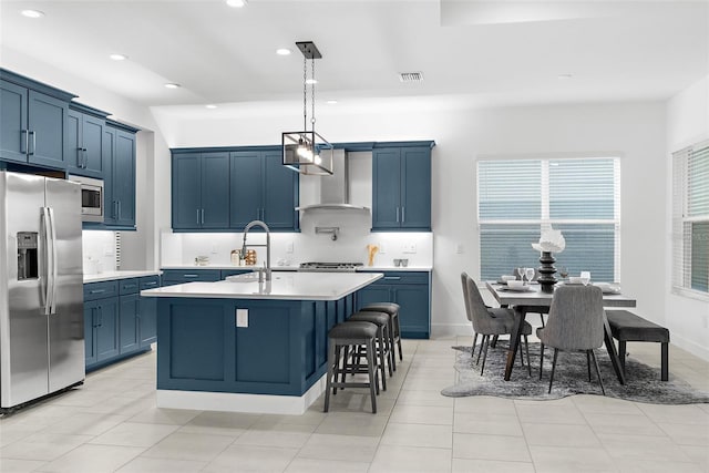 kitchen featuring stainless steel appliances, blue cabinets, an island with sink, hanging light fixtures, and wall chimney range hood