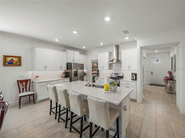 kitchen with a kitchen bar, appliances with stainless steel finishes, a kitchen island with sink, wall chimney range hood, and white cabinetry