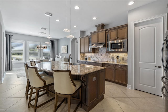 kitchen with light stone counters, a kitchen island with sink, sink, decorative light fixtures, and stainless steel microwave