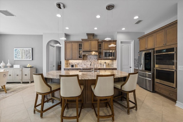 kitchen with pendant lighting, a center island with sink, sink, light stone countertops, and appliances with stainless steel finishes