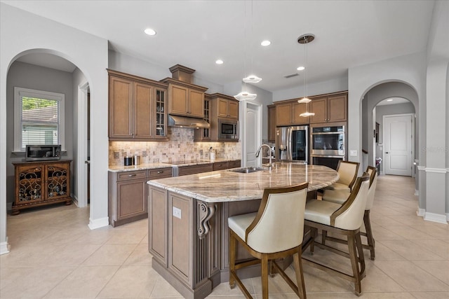 kitchen featuring a kitchen island with sink, hanging light fixtures, sink, light stone counters, and stainless steel appliances