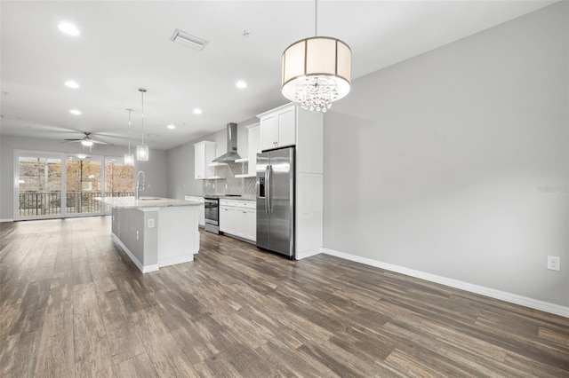 kitchen with white cabinets, wall chimney exhaust hood, an island with sink, appliances with stainless steel finishes, and decorative light fixtures