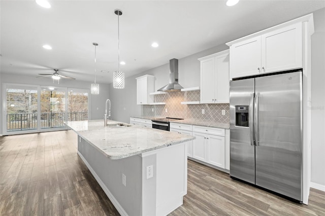 kitchen with hanging light fixtures, stainless steel appliances, wall chimney range hood, a center island with sink, and white cabinets