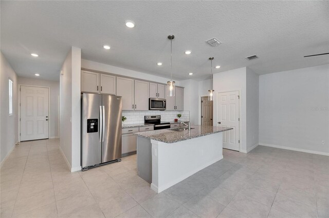 kitchen with stainless steel appliances, backsplash, dark stone countertops, decorative light fixtures, and a kitchen island with sink