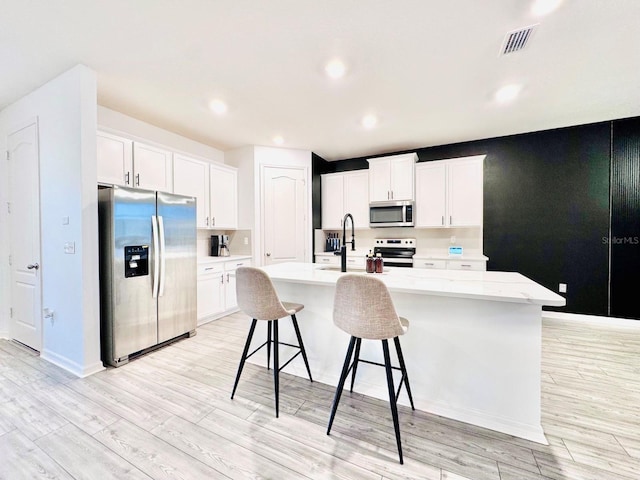 kitchen with light wood-type flooring, light stone counters, stainless steel appliances, a kitchen island with sink, and white cabinetry