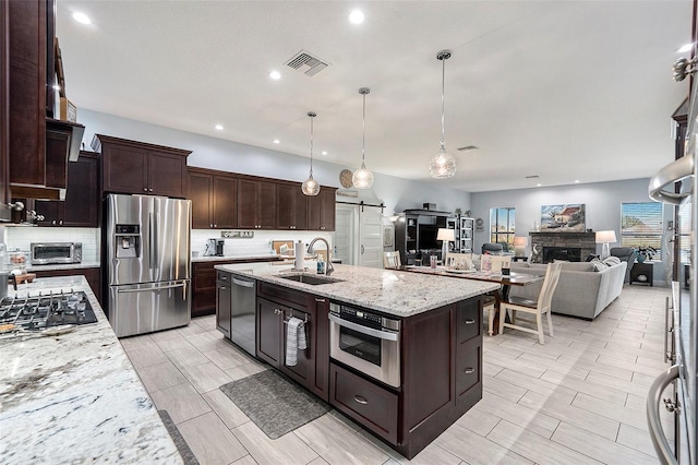 kitchen with a kitchen island with sink, sink, hanging light fixtures, light stone counters, and stainless steel appliances