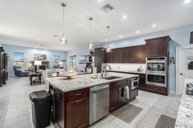 kitchen with a stone fireplace, hanging light fixtures, sink, an island with sink, and appliances with stainless steel finishes