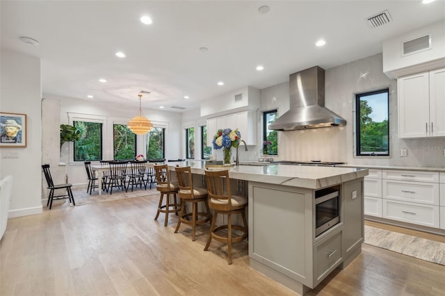 kitchen with stainless steel appliances, wall chimney range hood, pendant lighting, a center island with sink, and white cabinetry