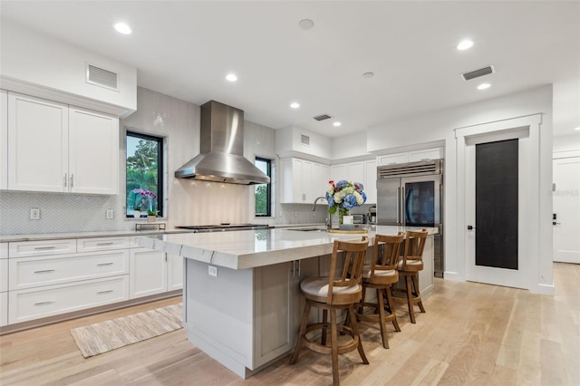 kitchen featuring white cabinetry, wall chimney exhaust hood, stainless steel appliances, light stone counters, and a center island with sink