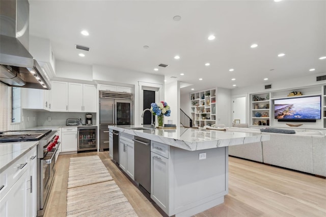 kitchen featuring high quality appliances, a center island with sink, white cabinets, wall chimney range hood, and wine cooler