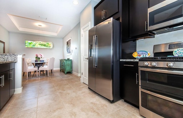 kitchen featuring stainless steel appliances, light stone counters, backsplash, a tray ceiling, and light tile patterned floors