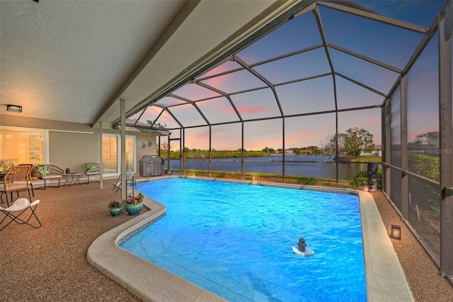 pool at dusk featuring an outdoor pool, glass enclosure, grilling area, a water view, and a patio area