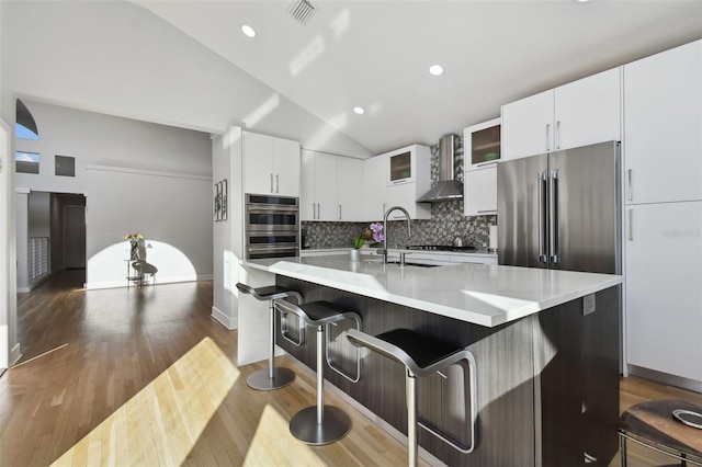 kitchen featuring backsplash, wall chimney range hood, vaulted ceiling, an island with sink, and white cabinetry