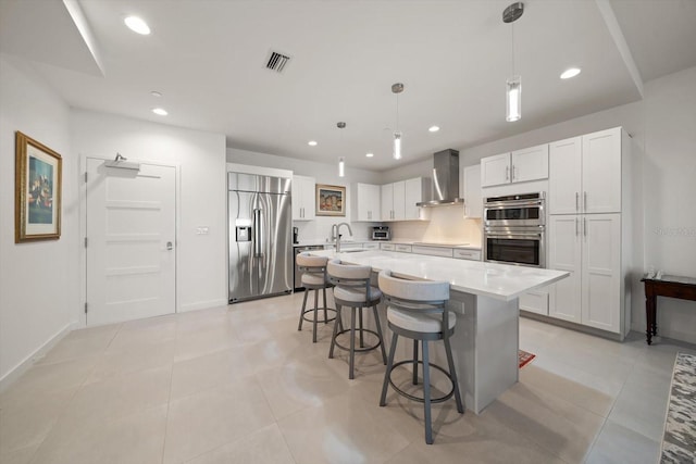 kitchen featuring a kitchen island with sink, wall chimney range hood, hanging light fixtures, appliances with stainless steel finishes, and white cabinetry