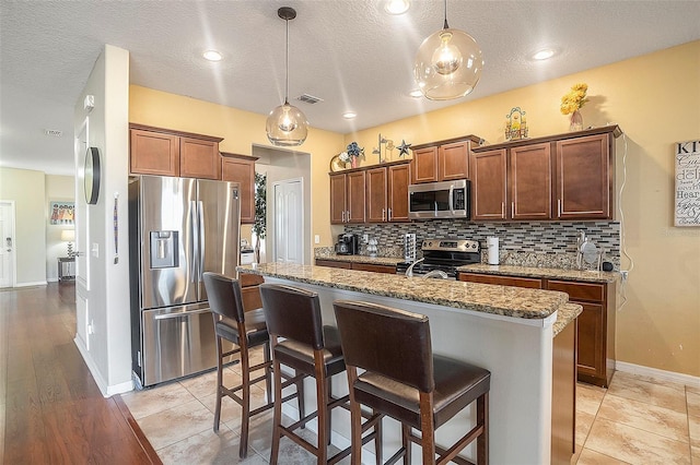 kitchen with light stone countertops, a center island with sink, hanging light fixtures, and appliances with stainless steel finishes