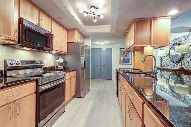 kitchen with sink, stainless steel appliances, dark stone counters, a tray ceiling, and light wood-type flooring