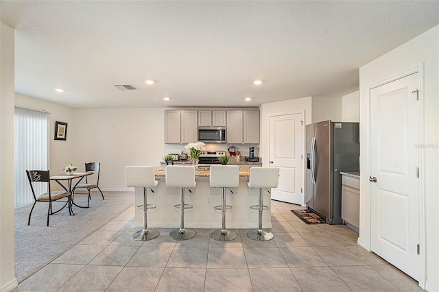 kitchen with an island with sink, light colored carpet, gray cabinetry, appliances with stainless steel finishes, and a kitchen breakfast bar