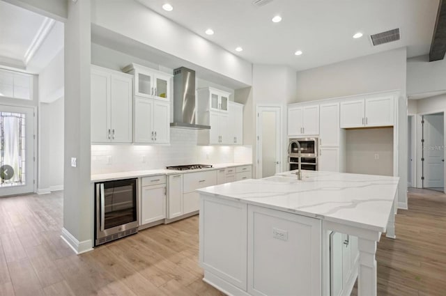 kitchen with white cabinetry, a kitchen island with sink, beverage cooler, and wall chimney range hood