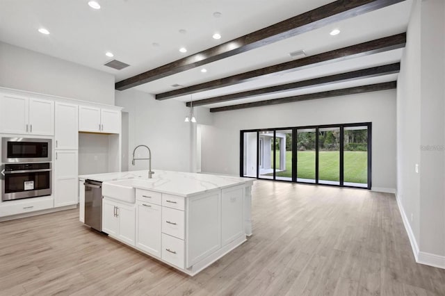 kitchen featuring beamed ceiling, appliances with stainless steel finishes, white cabinetry, light stone counters, and a center island with sink