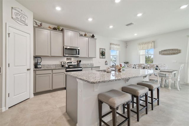 kitchen with gray cabinetry, a breakfast bar, a center island with sink, light stone countertops, and appliances with stainless steel finishes