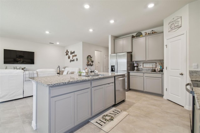 kitchen featuring gray cabinetry, sink, stainless steel dishwasher, an island with sink, and light stone counters