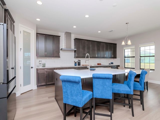 kitchen featuring wall chimney exhaust hood, pendant lighting, a center island with sink, a notable chandelier, and stainless steel refrigerator