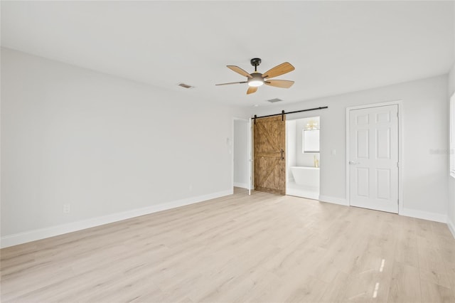 unfurnished bedroom featuring ceiling fan, a barn door, light wood-type flooring, and ensuite bath